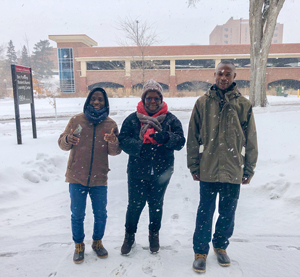 Three Cochran Fellows standing outside in the University campus while snow falls around them. Fellows are looking at the camera and smiling.